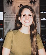 Lana Del Rey, with light-brown hair in an olive-colored shirt, smiling towards the camera at a fan meet