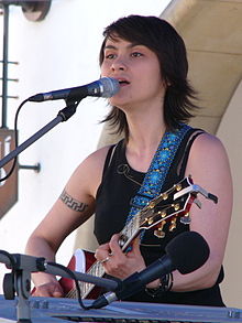 Lenka Dusilová standing onstage playing guitar, signing into a microphone