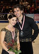 Waist high portrait of young dark haired woman and young dark haired man, both smiling, woman carrying red flowers