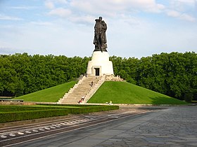 A large statue of a soldier holding a child and a sword, standing on a tall pedestal atop a grass-covered hill with steps leading up to the monument. Trees surround the background, and there are a few people sitting or walking on the steps.