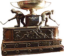 Two-handled metal cup with ice hockey players on each side on a wood base which has silver badges listing the winners