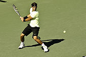 A brown-haired male tennis player with black shorts, a green shirt and a black headband swings a left-handed backhand on a hard court surface