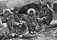 An Inuit family is sitting on a log outside their tent. The parents are engaged in domestic tasks. Between them sits a toddler. On the mother's back is a baby in a papoose. For infants and toddlers, the "set-goal" of the attachment behavioural system is to maintain or achieve proximity to attachment figures, usually the parents.