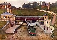 A steam train pushes a train through a station. A footbridge spans across the tracks from the station buildings at each side and tunnels are visible in the distance