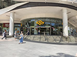 Photograph of station entrance at ground level. A curved transparent façade with stairs leading to the entrance