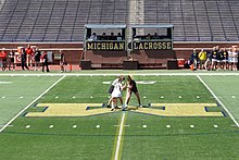 Two women, slightly hunched forward to each other, one in a black sleeveless top and short skirt and the other in a white version of the same clothing, stand in the middle of an artificial turf field with markings for gridiron football, while a third woman wearing a vertically black and white striped shirt stands just behind them with one hand between them, seen from some distance and above