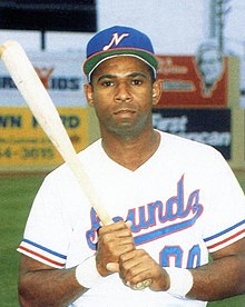 A man wearing a white baseball uniform with "Sounds" on the chest in blue and red and a blue cap with a white "N" poses holding a baseball bat with both hands.
