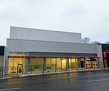 Photo of a light rail station entrance under construction which appears largely completed. It is a white building with glass panelling and an orange accent.
