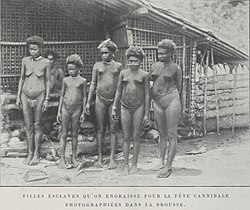 Black-and-white photograph of five nearly naked young women and girls standing in front of a hut; a sixth is visible in the background