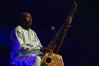 Photograph of Sissoko, seated onstage in a grand boubou robe, playing kora