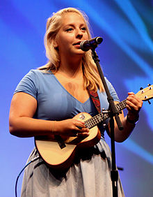 Julia Nunes wearing a light blue top and gray skirt, standing onstage with a ukulele, singing into a microphone