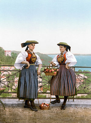 A photomechanical print of two middle aged women, standing on a hill or balcony. Both woman wear old colorful costumes with breast plates, skirts, and hats. The background below shows a waterbody and some houses.