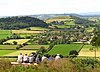 A patchwork of green and yellow fields with the roofs of houses visible centrally. In the background are hills and in the foreground the backs of several seated people.