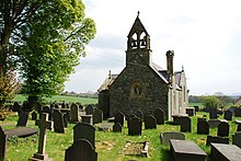 A small single-story church with bells hanging from a small stack, in a graveyard.