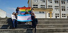 a group on the bleachers of a school football field, holding the rainbow and trans flags, symbolizing support for the LGBTI+ community.