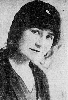 Portrait of a woman in a black cloche hat wearing a v-necked blouse under a dark jacket.