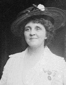 A smiling white woman with curly hair. She is wearing a hat with a wide brim, a light colored jacket, and medals pinned to her jacket.