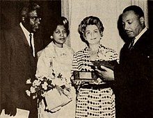 Photograph of two men flanking two women, one of whom holds flowers and the other holds a plaque.