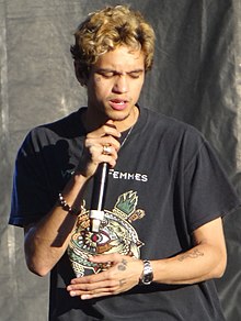 A young man with dark roots to his short bleached hair uses a cordless handheld mic to perform while wearing a black t-shirt with a colorful decal.