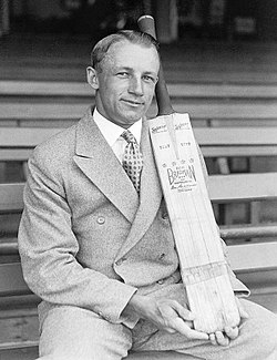 Cricketer Donald Bradman sits on a bench in a sports stadium and displays a cricket bat with his brand name on it. He wears a 1940s-style double-breasted suit and has his hair parted and slicked back with haircream, and is smiling.