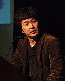 一名站在讲台上，身着棕色夹克和深灰衬衫的日本男子
