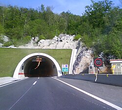 A tunnel portal with variable traffic signs indicating traffic flow direction and speed limit enforced are visible at the tunnel entrance and to the side of the road