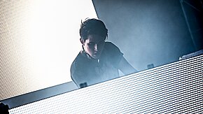 Porter Robinson stands at a table with a brightly lit screen; another screen is behind him