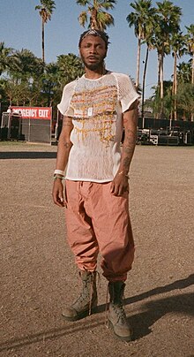 Hendricks stands in front of palm trees and stage equipment in the bright sun, wearing sturdy boots.