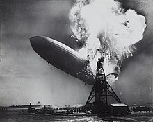 A black and white photograph of an airship near a mooring mast exploding at its stern.