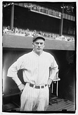 A man in a white baseball uniform stands in front of a dugout with his hands on his hips.