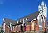 A brick church, seen uphill from a corner, with a tall white steeple. One of its front windows has been boarded up.