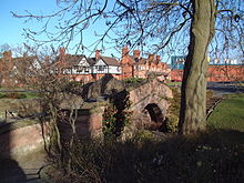 A small arched sandstone bridge seen from an angle, with a tree to the right and houses and factory buildings in the background.