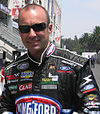 A man in his early thirties wearing black rectangular sunglasses and racing overalls with sponsors logos. He is smiling at the camera