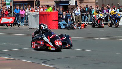 Sanctioned motorsport photographers identified by individually numbered white bibs on a road-course in the Isle of Man with public spectator area behind.