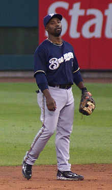 A man wearing gray pants, a navy blue jersey with "Brewers" written in white, a navy blue cap bearing a white "M", and a baseball glove stands on a baseball field.
