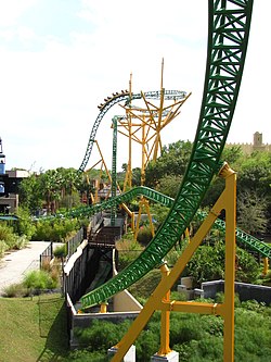 A vertical photograph of Cheetah Hunt's layout from the Sky Ride gondola. Pictured is the yellow colored supported windcatcher tower with green track where a Cheetah Hunt train ascends. The photograph features the descending track into a trench and over the Sky Ride, as well as the last bunny hill of the layout.