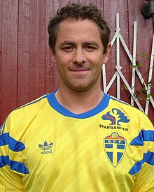 A head and chest photo of Ken Burvall, a white man in his thirties with short brown hair and no beard. He is wearing a yellow football jersey with blue accents and is standing in front of a wooden wall, painted red.