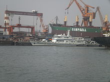 patrol boat berthed among tankers in a shipyard, gantry cranes in the background