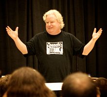 A man in giving a presentation to an audience in front of a theatrical curtain