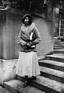 Lavaud in a hat and coat standing on the steps of a building.