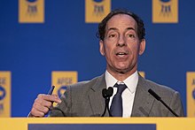 An older man with curly black hair speaking in front of an indoor lectern in front of a blue and yellow backdrop