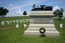 A white marble monument shaped like an altar with steps, with on top a bronze model of a steam engine, on a grassy cemetery with white headstones in the background