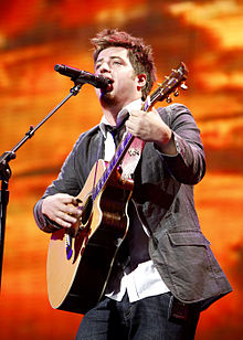 A Caucasian man sings into a microphone while playing a guitar.