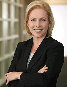 A wide portrait shot of a smiling, middle-aged Caucasian female (Kirsten Gillibrand) looking straight ahead with arms crossed. She has long blonde hair, and is wearing a dark blazer with a white top. She is placed in front of a light background.