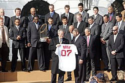 Two rows of men in suits, one holding a large trophy, stand while a smiling man stands in front of them holding a white baseball jersey which reads "BUSH 07" on the back.