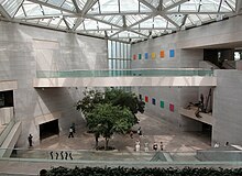 The interior of the National Gallery East Building. A large open cement room contains several people on the ground far below a balcony. Several trees are planted in the concrete floor, and an array of clear windows let in sunshine from above.