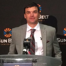 A color photograph of Neal Brown in a suit and tie at a podium.