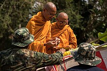 Two Buddhist Monks in Saffron robes with shaved heads and amber-brown skin stand over a pillar adorned with yellow flora. Two soldiers in green camo and bucket hats present the pillar to the monks.