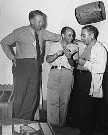 Three men talking. The one on the left is wearing a tie and leans against a wall. He stands head and shoulders above the other two. The one in the center is smiling, and wearing an open-necked shirt. The one on the right wears a shirt and lab coat. All three have photo ID passes.