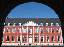 Through the archway of a gate is visible a wide part of the red-brick façade of a two-storey building, with large windows outlines with white stone arches. They grey roof shows skylight windows of a third floor. The entrance is preceded by a small set of steps and is surmounted by an ornament showing two figures supporting a coat of arms.